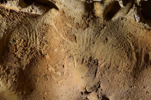 Eine ockergelbe Höhlenfelswand mit zwei Objekten aus gebogenen Linien und weitere Gravuren