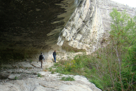 Ein grauer Steinweg im Gebirge, darüber ein mächtiger Felsüberhang, rechts grüne Bäume und Büsche